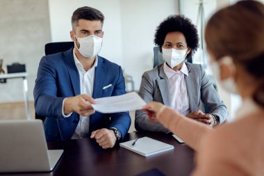 Members of human resource team wearing protective face masks while taking CV from a candidate during job interview in the office. 