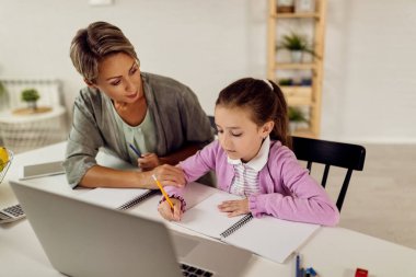 Little girl taking notes while e-learning over laptop with help of her mother at home. 