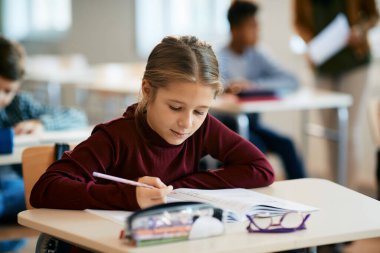 Elementary student writing at her desk during a lesson in the classroom.