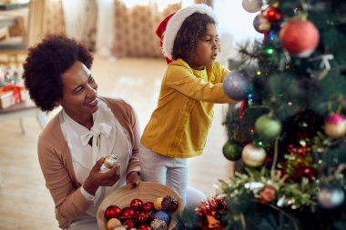 Happy African American mother enjoying in decorating Christmas tree with her daughter at home. 