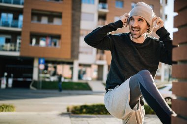 Young happy sportsman adjusting his headphones while stretching and warming up on the street. 