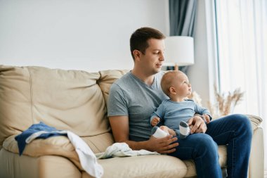 Young smiling father sitting on the sofa with his baby boy while spending time with him at home. 