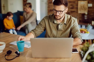 Male entrepreneur  using computer and having a cup of coffee while working at home. His wife and daughter are in the background. 