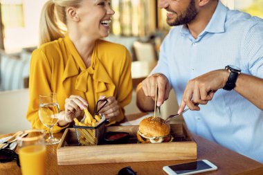 Close-up of happy couple eating French fries and cheeseburger during lunch in a restaurant.
