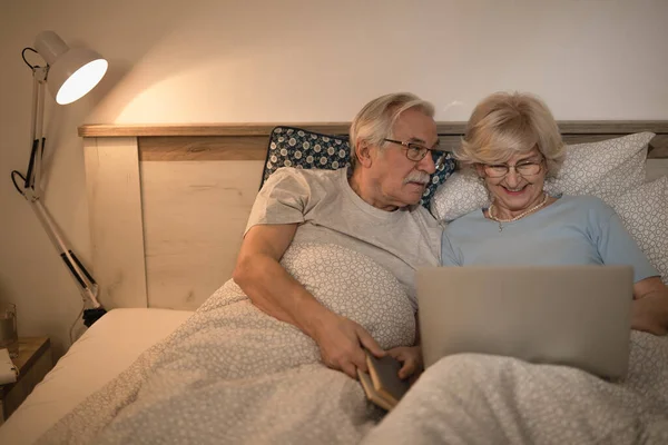 Senior man lying down in bed and talking to his wife who is surfing the net on laptop.