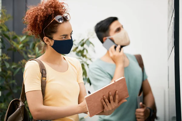 College student wearing face mask and writing in her notebook while reading notifications on bulletin board in a hallway.