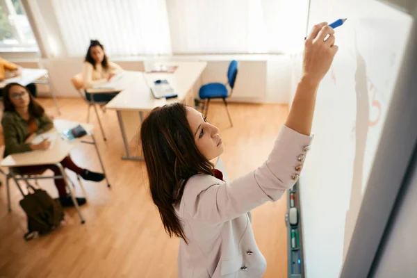 Female professor writing on smart board while giving lecture to her students in the classroom.