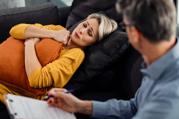 Distraught woman talking to a mental health professional while lying down on psychiatrist's couch and hugging a pillow.