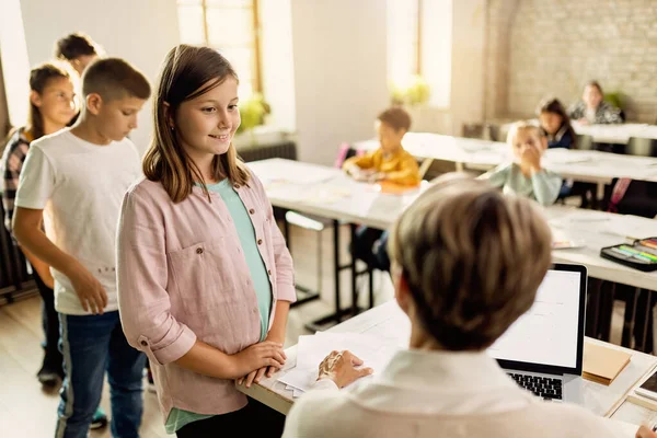 Happy little girl talking with teacher while her classmates are waiting in the line behind her in the classroom.