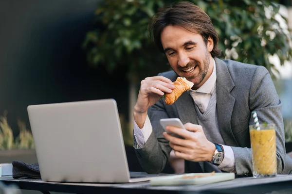 Young Smiling Entrepreneur Eating Croissant Using Smart Phone While Having — Photo