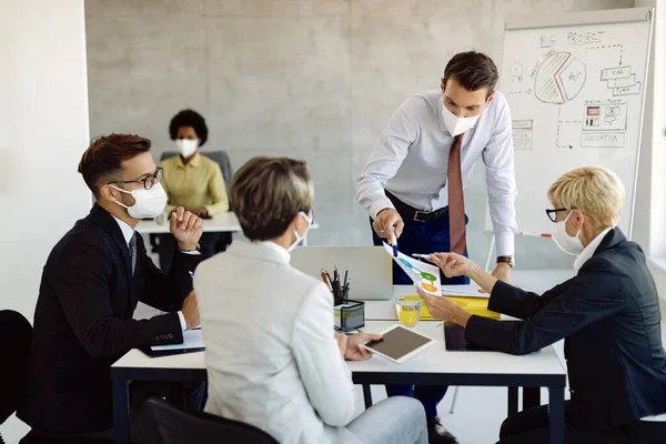 Group of entrepreneurs with protective face masks brainstorming while analyzing business graphs on a meeting in the office.