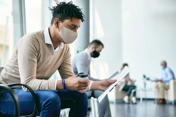 Young black businessman with face mask analyzing his CV while sitting in waiting room and preparing for job interview.