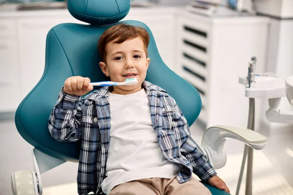 Small kid sitting in dentist\'s chair and learning the proper way of brushing teeth at dentist\'s office.