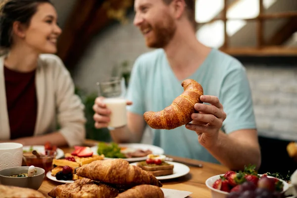 Close-up of man eating croissant while having breakfast with his girlfriend at home.
