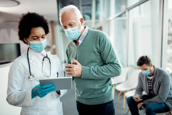 African American female doctor and her senior patient using digital tablet while wearing protective face mask at hospital hallway.
