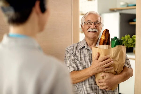 Happy senior man standing at doorway  and holding bag of groceries delivered by a courier.