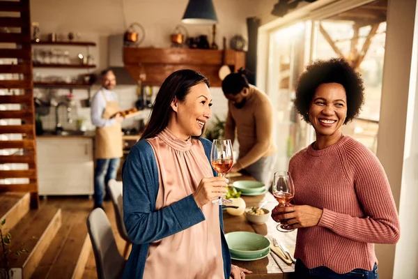 Happy African American woman and her Asian female friend drinking wine while their husbands are preparing dinner in the background.