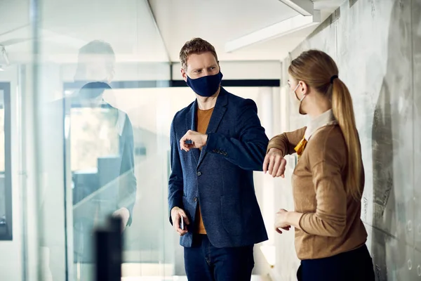 Happy business colleagues with face masks elbow bumping while greeting in hallway of an office building.