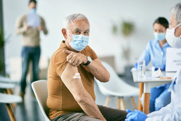 Mature man with face mask getting vaccinated against coronavirus and showing adhesive bandage on his shoulder at vaccination center.