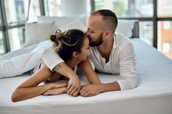 Young couple in love lying down in bed. Man is kissing his girlfriend on forehead.