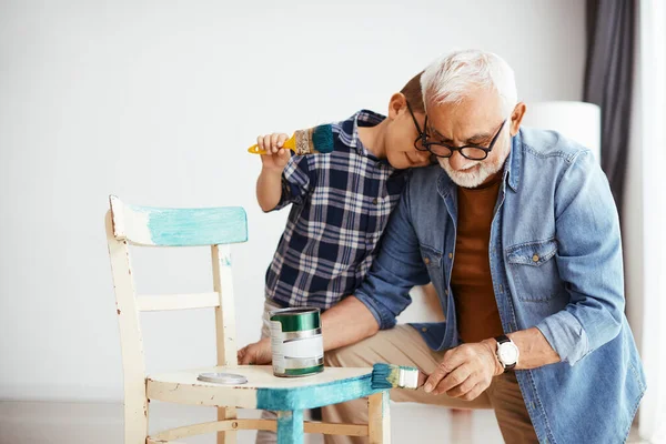 Senior man painting wooden chair while grandson is embracing him at home.
