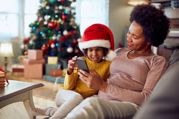 Happy black mother and her small daughter text messaging on mobile phone while relaxing on the sofa during Christmas day.