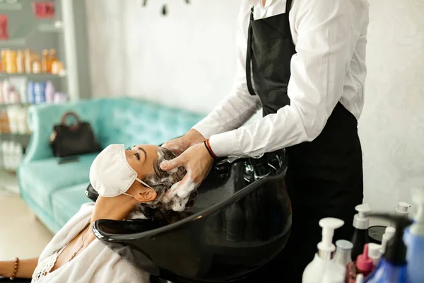Happy woman with face mask enjoying while hairdresser is washing her hair at the salon during coronavirus epidemic.