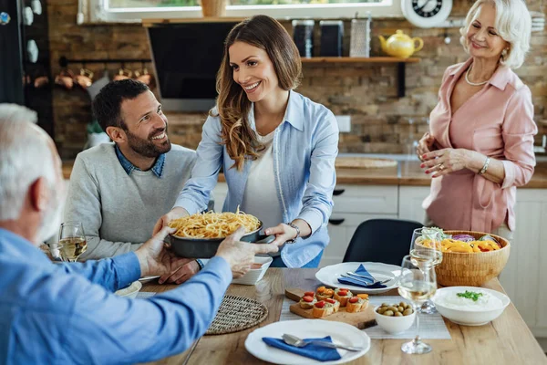 Young happy couple having family lunch with senior parents in dining room. Focus is on woman serving food at the table.