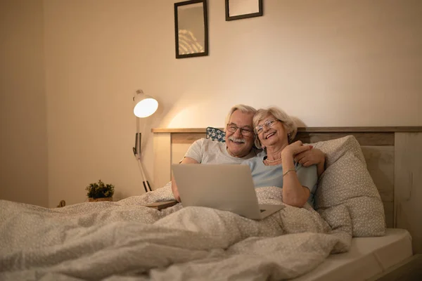 Happy senior couple lying down in bed and surfing the net on laptop at night.