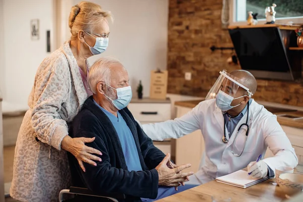 Mature couple and black doctor wearing protective face masks while talking at nursing home.
