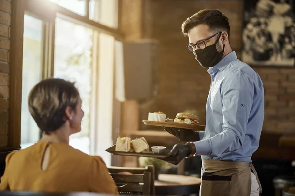 Happy waiter serving food to a guest while wearing protective face mask and gloves in a pub.