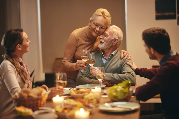 Embraced senior couple celebrating marriage anniversary and toasting with wine during family meal at dining table.