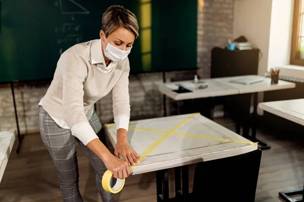 Female teacher with face mask marking empty tables with a tape in the classroom during COVID-19 pandemic.