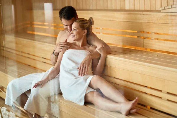 Happy couple talking while sitting close to each other in sauna at health spa.  The view is through the glass.
