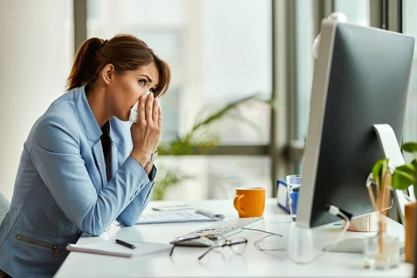 Young entrepreneur sneezing in a tissue while working at her office desk.