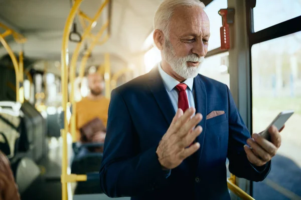 Happy mature businessman having video call over smart phone while commuting by public transport.