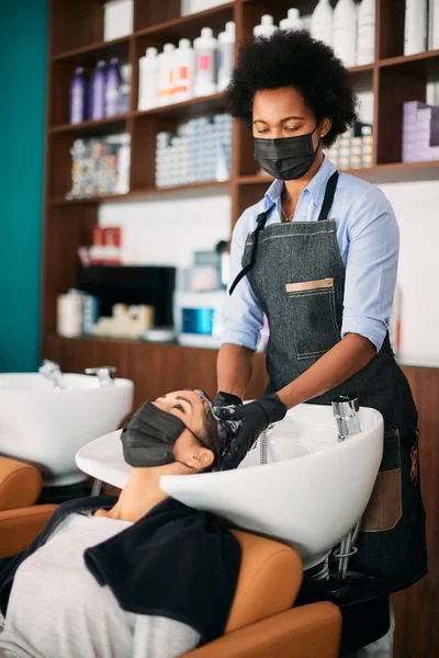 Black female hairdresser wearing protective face mask while having hair woman\'s hair at the salon.