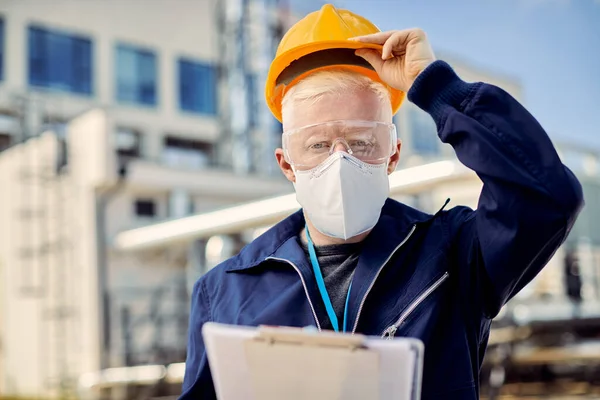 Civil engineer with protective face mask analyzing paperwork and looking at camera at construction site during COVID-19 epidemic.