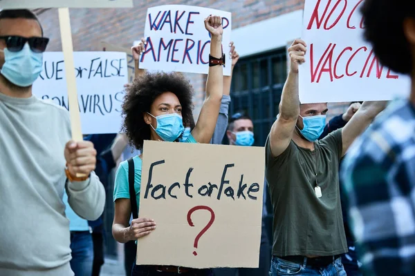 African American woman with raised fist wearing protective face mask while carrying banner with \'fact or fake\' inscription during public demonstrations.