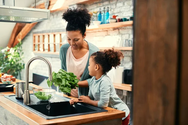 Happy black mother and her small daughter washing salad while preparing food in the kitchen.