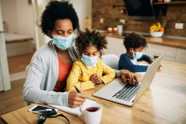 African American working mother using laptop and taking notes while daughter is sitting in her lap at home during COVID-19 pandemic.