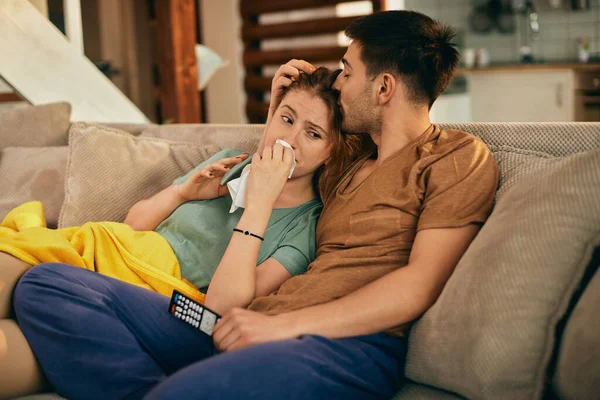 Young caring man kissing his girlfriend who is crying while watching sad movie on TV at home.