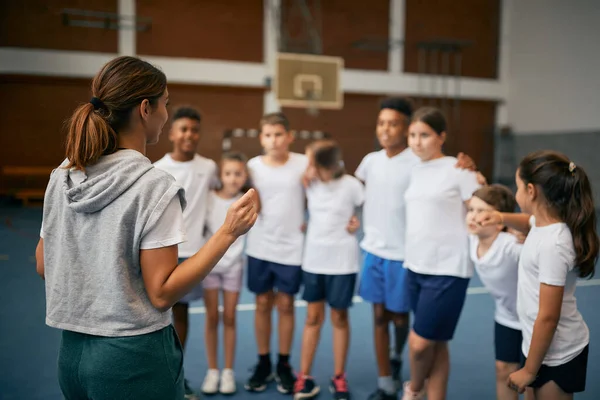 Back view of female PE teacher explaining exercise activities to group of kids at school gym