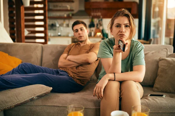 Displeased woman changing channels and feeling uncertain what to watch on a TV with her boyfriend at home.