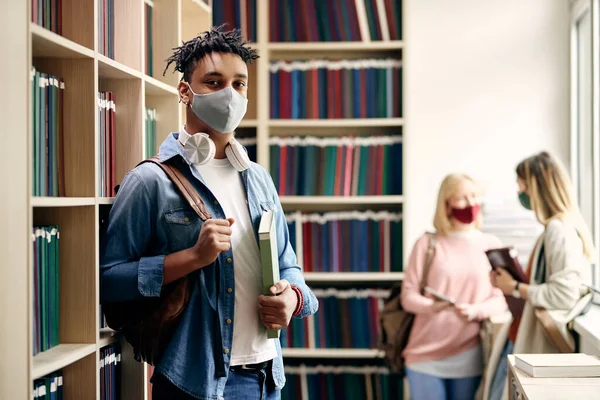 Portrait of smiling black student wearing protective face mask while studying in library during coronavirus pandemic. There are students in the background.