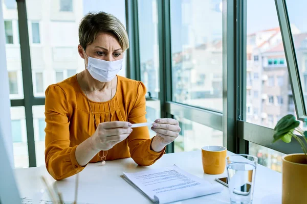 Businesswoman with face mask using thermometer while measuring her body temperature in the office.