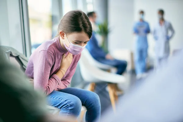 Small girl feeling ill and holding her chest in pain while sitting in waiting room at medical clinic during coronavirus pandemic.