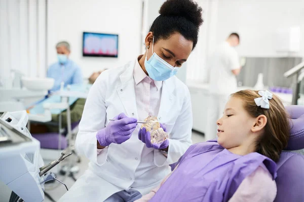 Black female dentist using artificial dentures while communicating with little girl during dental exam.