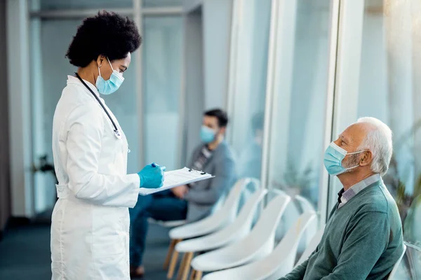 Black female doctor going through medical record while being with her senior at hospital waiting room. Both of them are wearing protective face masks.