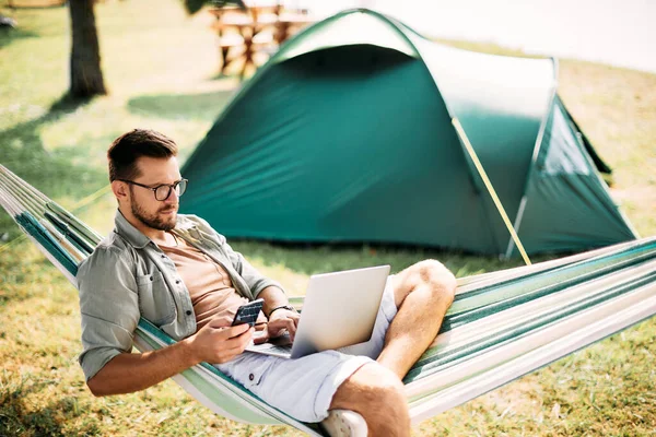 Male entrepreneur working on laptop and texting on mobile phone while being on camping in nature.